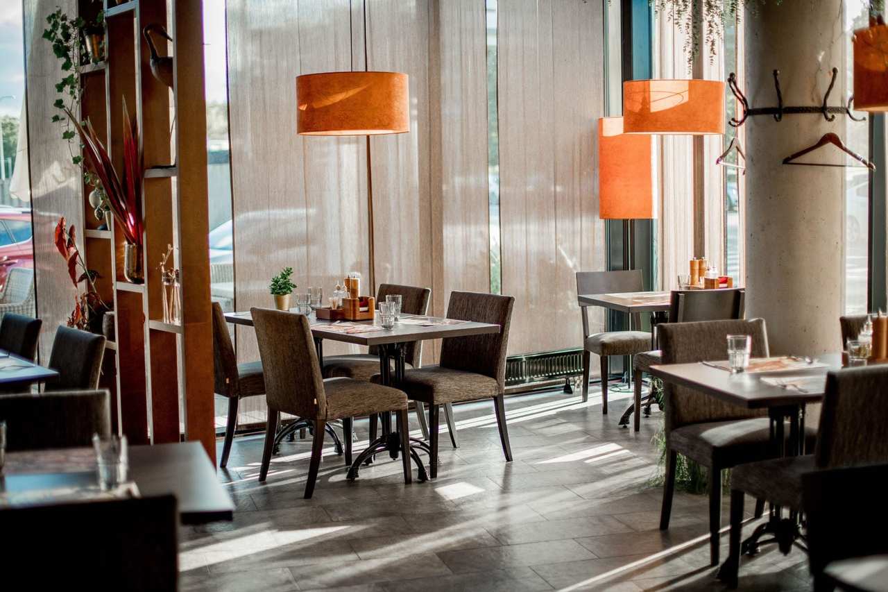 Comedor vacío con luz de tarde, mesas preparadas con vasos y lámparas naranjas.