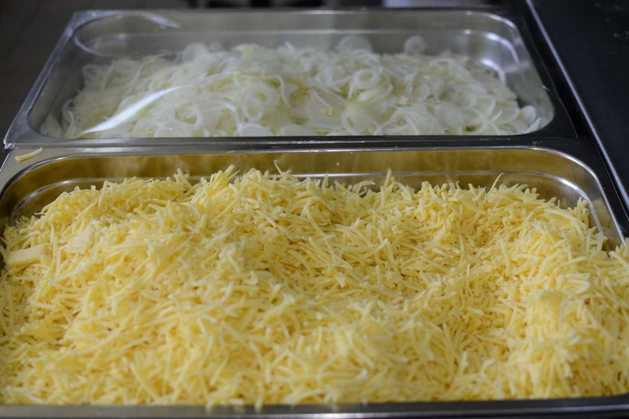 Dos bandejas de acero sobre una mesa de cocina: una con cebolla en juliana y otra con queso rallado.