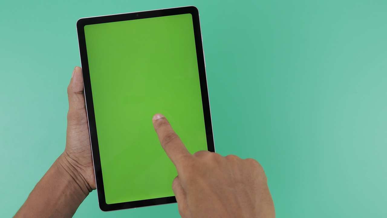 Una mano sostiene una tableta con la pantalla en verde liso y la otra señala la pantalla con un dedo.